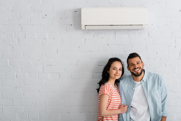 Junges Paar lächelt in die Kamera, während es in der Nähe einer Klimaanlage an der Hauswand steht  - Foto, Bild