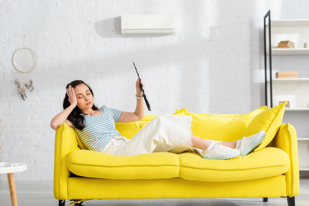Selective focus of tired girl holding fan while suffering from heat on sofa in living room - 写真・画像
