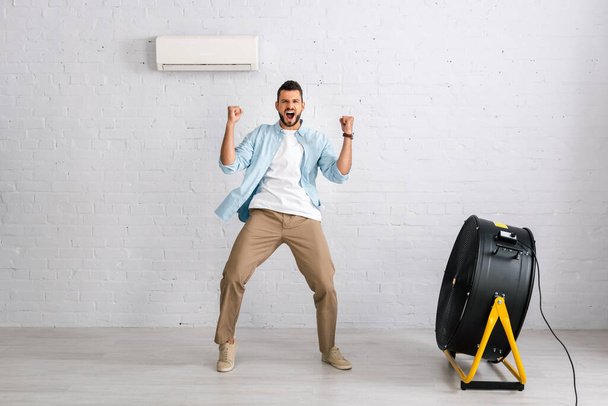 Positive man showing yes gesture while standing near air conditioner and electric fan in living room - Foto, Bild