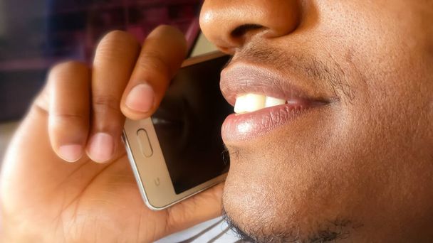 Handsome young man on a phone call. Listening. Close up. Side view. Attentive and smiling with his white teeth showing. African man with light skin. Holding, using a smartphone with one hand to his ear. Partial view of his face. - Foto, imagen