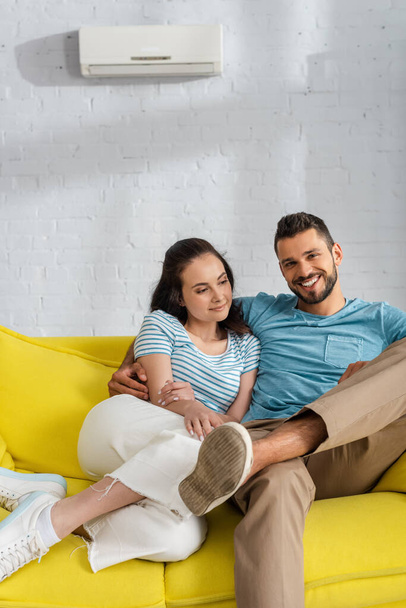 Handsome man smiling at camera while hugging attractive girlfriend on couch in living room - Фото, изображение