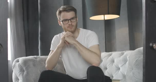 Portrait of brunette man taking off eyeglasses and looking at camera with serious facial expression. Middle shot of bearded Caucasian guy sitting on couch indoors and thinking. Cinema 4k ProRes HQ. - Felvétel, videó