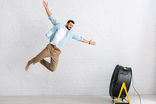 Handsome bearded man jumping near electric fan at home  - Fotó, kép