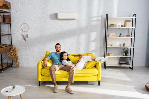 Hezký muž se usmívá na kameru v blízkosti krásné přítelkyně ležící na gauči v obývacím pokoji - Fotografie, Obrázek