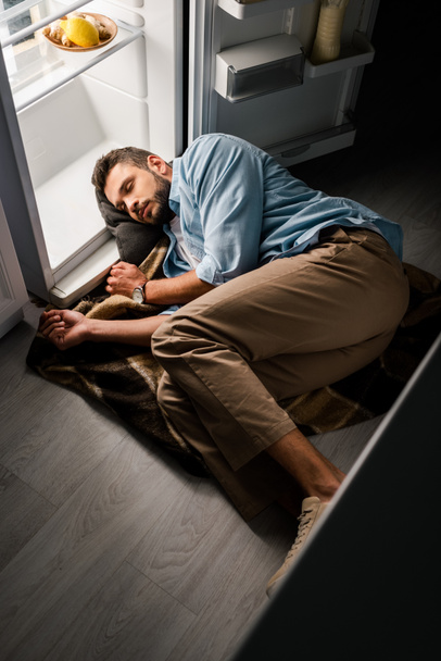 Vue grand angle de l'homme dormant près d'un réfrigérateur ouvert sur le sol la nuit  - Photo, image