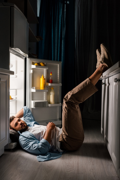 Zijaanzicht van de mens kijkend naar de camera terwijl hij op de vloer ligt in de buurt van een open koelkast 's nachts  - Foto, afbeelding