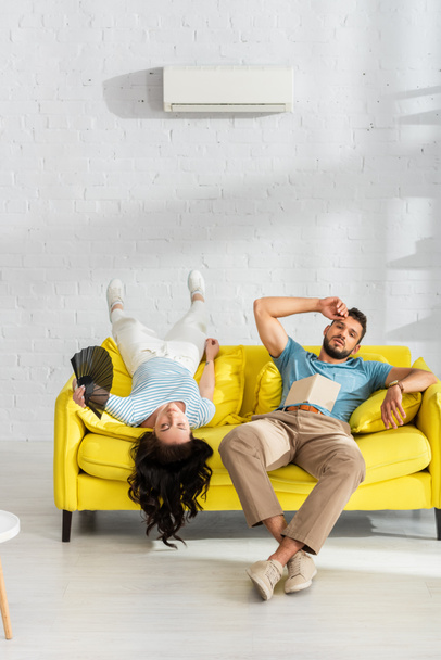 Müdes Paar mit Buch und Ventilator leidet unter Hitze auf Sofa im Wohnzimmer - Foto, Bild