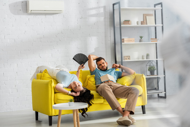 Selektiver Fokus eines erschöpften Paares mit Ventilator und Buch, das sich im Wohnzimmer heiß fühlt - Foto, Bild