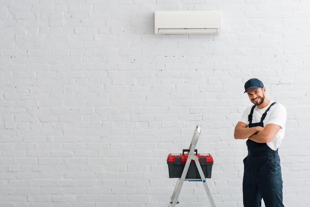 Glimlachende werkman met gekruiste armen kijkend naar camera nabij gereedschapskist op ladder en airconditioner aan muur - Foto, afbeelding