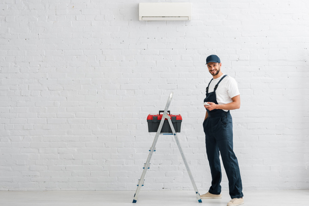 Obrero sonriente sosteniendo controlador remoto de aire acondicionado cerca de la caja de herramientas en la escalera
  - Foto, Imagen