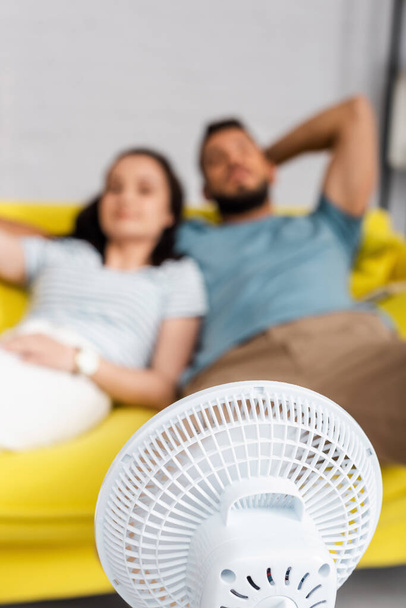 Enfoque selectivo de ventilador eléctrico cerca de pareja sentada en sofá en casa
  - Foto, imagen