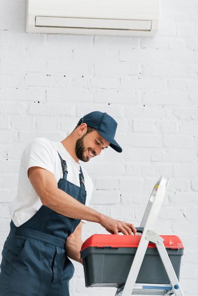 Trabajador sonriente en caja de herramientas de sujeción uniforme cerca de escalera y aire acondicionado en pared blanca
 - Foto, imagen