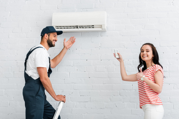 Side view of smiling woman with remote controller of air conditioner showing thumb up near workman in overalls on ladder  - Foto, Imagem