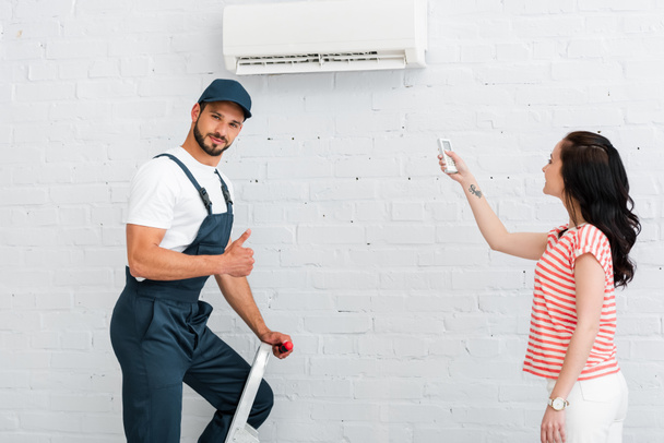 Komea työmies osoittaa peukalo ylös lähellä hymyilevä nainen kauko-ohjain ilmastointilaite  - Valokuva, kuva