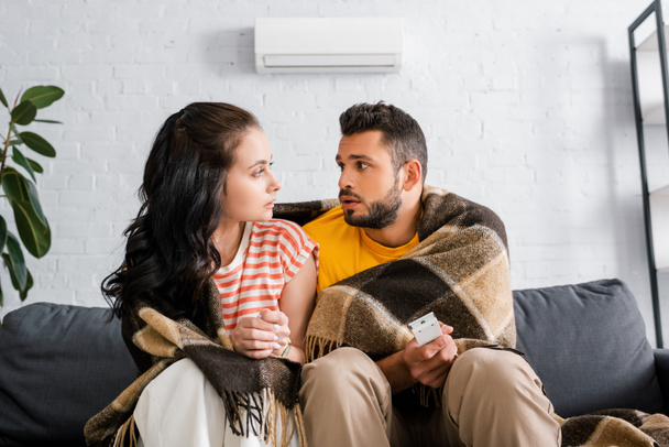 Young woman looking at boyfriend wrapped in plaid and holding remote controller of air conditioner on couch  - Foto, Imagem