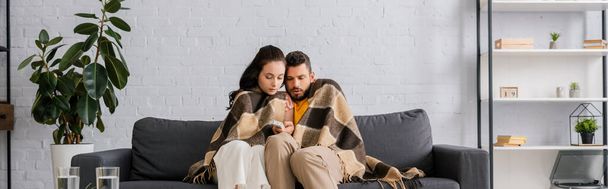 Panoramic crop of couple wrapped in plaid holding remote controller of air conditioner on sofa  - Photo, Image
