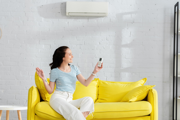 Smiling woman holding remote controller of air conditioner on couch at home  - Fotoğraf, Görsel