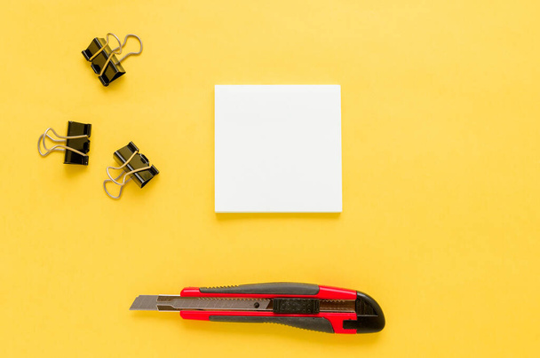 White square notes, red office knife, bulldog paper clip on yellow background with copy space. Mock up. Top view. Concept chancellery, office supplies, accessories - Fotoğraf, Görsel