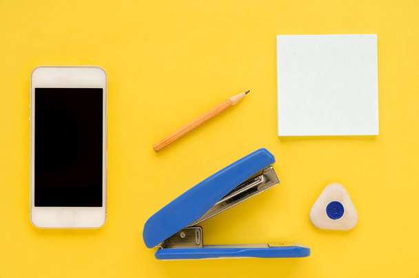 White square notes, pencil, eraser, blue stapler, sharpener, white smartphone on yellow paper backgroun - Zdjęcie, obraz