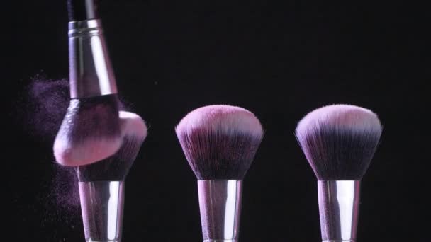 Concepto de belleza. Cepillos cosméticos con polvo cosmético rosa extendiéndose sobre fondo negro en cámara lenta
 - Metraje, vídeo