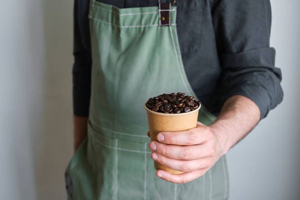 A paper cup with coffee beans in the hands of a barista. Ready to brew roasted coffee. A man in a culinary apron. Fresh organic coffee. Disposable paper cup. Making coffee, professional service - Photo, image