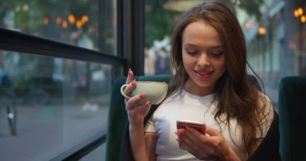 Közelről lőtték le. A lány egy kávézóban ül a verandán. Teát iszik és sms-ezik telefonon. A mosolygós lány takaróba van csomagolva. 4K - Felvétel, videó