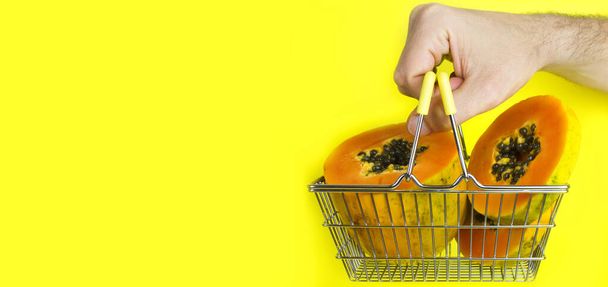 Cesta de compras con papaya de frutas tropicales y mano de hombre sosteniendo la cesta sobre fondo amarillo. Compras en el supermercado. Concepto de comida saludable. Compras en línea. Comida fresca y entrega de fruta. Espacio para el texto. Banner
 - Foto, imagen