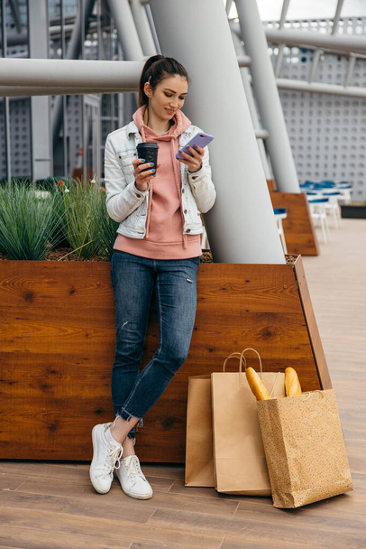 Lächelnde junge Frau nutzt Smartphone, um nach dem Einkauf ein Taxi zu nehmen, Kaffeetasse in der Hand, Einkaufstasche mit langem weißem Baguette über dem Hintergrund der Stadt - Foto, Bild