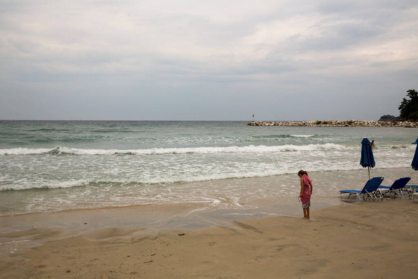 Dziewczyna w różowym kolorze stojąca przy piaszczystej plaży i mocząca nogi w morzu podczas burzliwej pogody latem - Zdjęcie, obraz