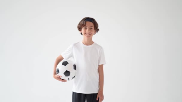 Fotbalista. Portrét roztomilého šťastného chlapce v bílém tričku držícího fotbalový míč v jedné ruce a usmívajícího se na kameru, zatímco stojí na šedém pozadí - Záběry, video