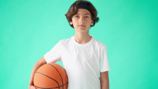 Fiatal kosaras. Portré aktív tinédzser fiú fehér pólóban kezében kosárlabda és mosolyog a kamera, szemben türkiz kék háttér - Felvétel, videó
