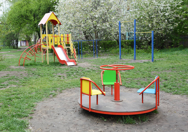 Üres szabadtéri játszótér csúszdával, körhintával, húzódzkodással, állkapcsokkal a parkban virágzó fákkal a háttérben. - Fotó, kép