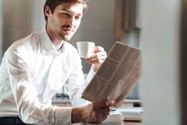 Photo of pleased brunette businessman wearing shirt drinking coffee and reading newspaper at modern kitchen - Фото, зображення