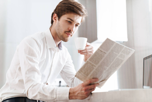 Photo of thinking brunette businessman wearing shirt drinking coffee and reading newspaper at modern kitchen - Fotó, kép