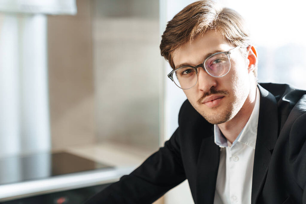 Photo of serious handsome businessman wearing eyeglasses posing and looking at camera in modern kitchen - Fotografie, Obrázek