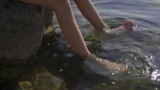 Портрет подростка, сидящего на большом камне и смочивающего ноги в воде. Мальчик держит в руке водолазную маску. Море, утро, замедленная съемка
. - Кадры, видео