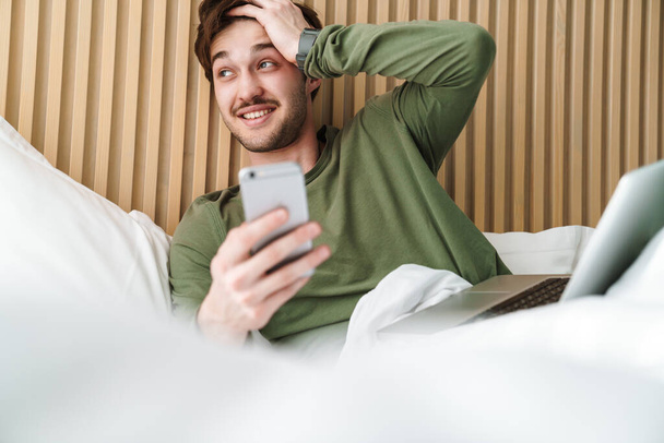 Fénykép kaukázusi mosolygós férfi bajusszal segítségével mobiltelefon és laptop, miközben pihen az ágyban - Fotó, kép