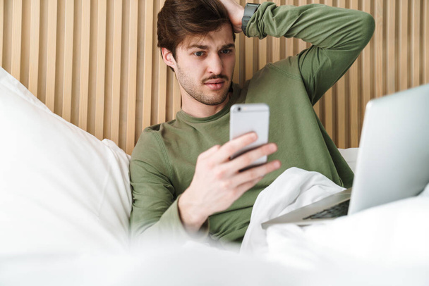 Zdjęcie kaukaskiego zdezorientowanego mężczyzny z wąsami za pomocą telefonu komórkowego i laptopa podczas odpoczynku na łóżku - Zdjęcie, obraz