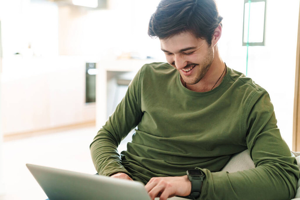 Photo of laughing young man with mustache typing on laptop while sitting on couch in apartment - Fotografie, Obrázek