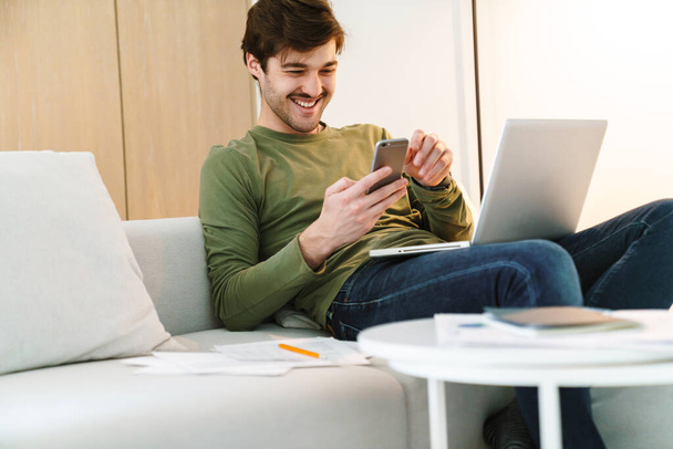 Photo of laughing young man with mustache using smartphone and laptop while sitting on couch in apartment - Foto, Imagem
