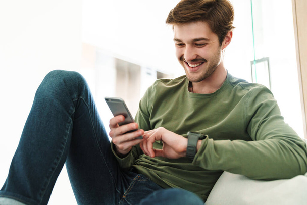 Photo of cheerful young man with mustache smiling and using smartphone while sitting on couch in apartment - Fotoğraf, Görsel