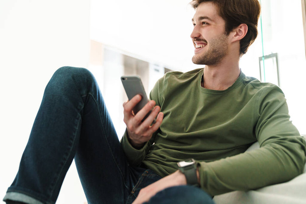 Photo of cheerful young man with mustache smiling and using smartphone while sitting on couch in apartment - Fotó, kép