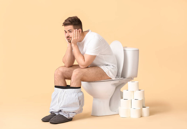Young man with rolls of paper sitting on toilet bowl against color background - Foto, imagen