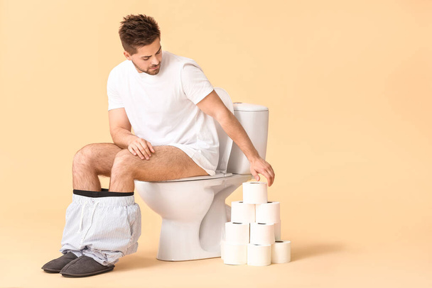 Young man with rolls of paper sitting on toilet bowl against color background - Foto, immagini
