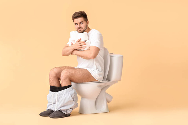 Jeune homme avec des rouleaux de papier assis sur un bol de toilette sur fond de couleur - Photo, image