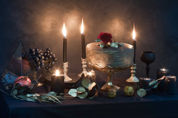 mysterious looking still life with handmade figure of cake, burning candles, dried rose and pomegranate in a prism, craft art of wedding decoration details - Photo, image
