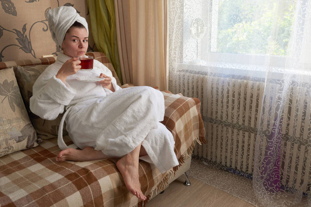 Egy fehér fürdőköpenyes lány pihen a kanapén, teázgat.. - Fotó, kép
