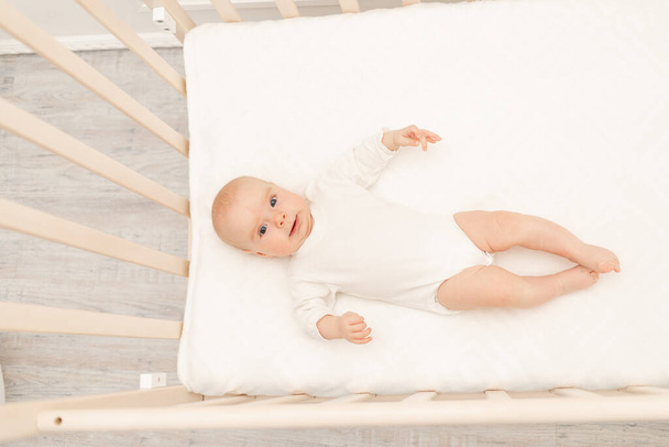 白いベビーベッドで眠っている赤ん坊と彼のおもちゃ。子供のための保育室のインテリアと寝具。かわいい男の子が寝そべってる。子供は白い寝室で昼寝をしている。ボディスーツのパジャマの健康な子供. - 写真・画像