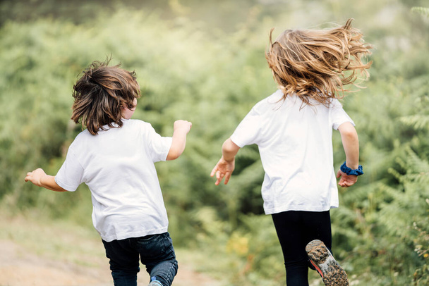 Twee broers een 4-jarige bruine jongen en een 7-jarig blond meisje rennen terug naar de camera door heel mooie groene bomen en geel zonlicht - Foto, afbeelding