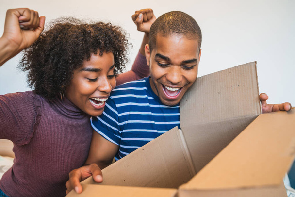 Portrait of happy young latin couple opening a package at home. Delivery, shipping and postal service concept. - Фото, изображение